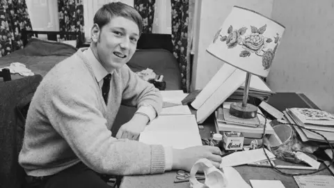John Downing/Getty Images A black and white photo in which a young man sits at a desk full of books.