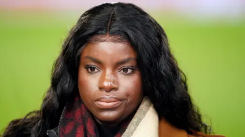 Eni Aluko, who has black wavy hair and wears a scarf over a brown leather coat, stands in front of a football pitch with a serious expression on her face. 