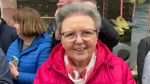 A woman wearing glasses and a pink coat stands in front of a small crowd of other people. She is wearing a pink scarff and has silver hair