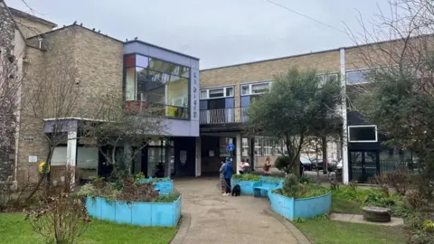 Great Yarmouth's central library, with a pathway, bordered by plants in "boat" shaped, blue raised beds, and community garden on the approach. The building features a large box window above the entrance. A few people are standing outside.