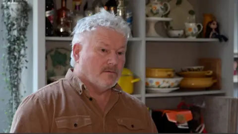 Shaun Whitmore/BBC A head and shoulders image of a man with white-grey short hair and a beard. He is wearing a brown shirt with black buttons.