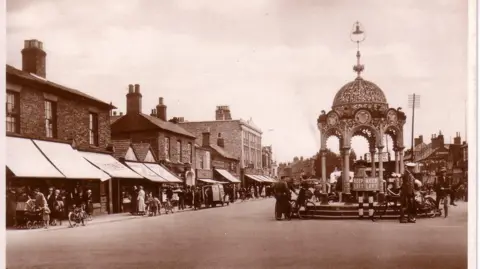 Black and white photo from 1934 showing a busy row of shops on the left, some with large canopies, on the right is the fountain monument, an ornate eight-sided structure with a domed roof, with what looks like a bell at the top of a rod extending vertically from the top