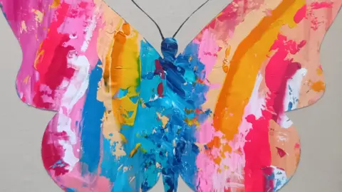 A butterfly-shaped artwork called My Angel by Allegra Dams is painted in bright pink, orange, yellow and blue colours against a plain background.