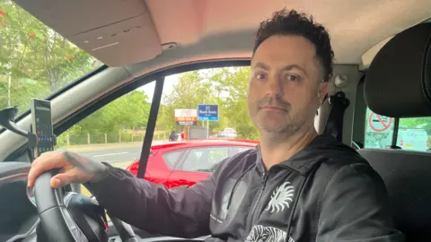 BBC Manuel Avac sat in the mini-bus wearing a black-hooded jacket. he has black curly hair and dark stubble. 