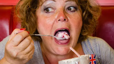 One of Emma's photographs. She pictures a woman with flowy ginger hair eating an ice cream with a long spoon. She is putting the ice cream in her mouth - whilst sitting on a red leather seat. A Union Jack flag sits limply out the top of her ice cream.