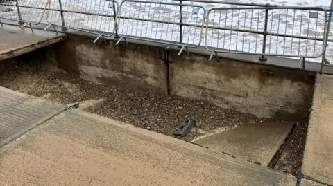 GUY CAMPBELL/BBC Metal fencing is in background and a large, rectangular-shaped hole in a concrete promenade has shingle inside and the remnants of the collapsed walkway.