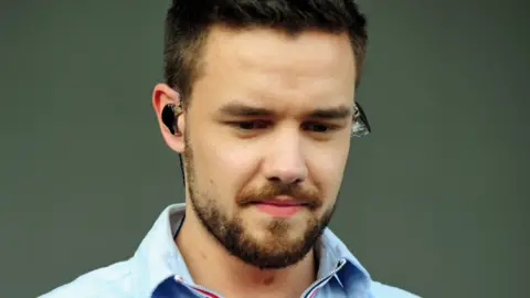 Liam Payne, with dark hair and a beard, is looking down and wearing an earpiece in front of a plain background.