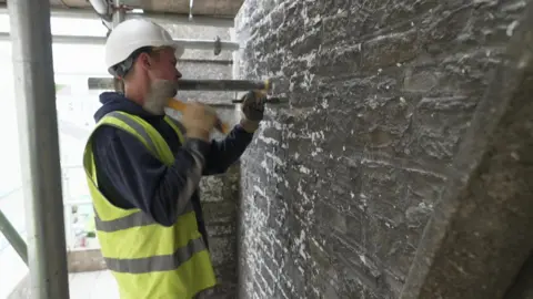 BBC A stonemason hits a chisel on the grey wall of a church in Devon. He is wearing a blue hoodie, yellow high-vis jacket and a white hard hat.
