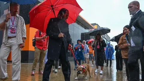 Getty Images Lewis Hamilton in black coat and trousers with his bulldog on a lead. He is holding a red umbrella to shield himself from the rain.