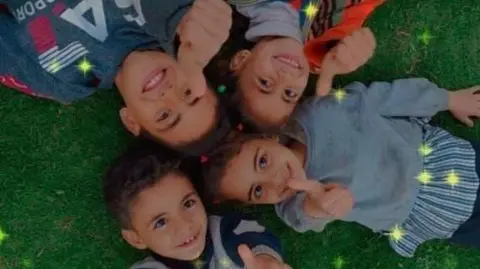 Handout Four children are lying in a circle on the grass with their thumbs in the air. Above the picture you can see some yellow stars that were added with a camera filter.