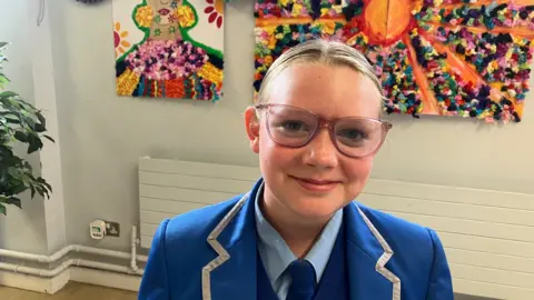 A schoolgirl is smiling into the camera. Her uniform is blue. She wears glasses and her hair is tied back.