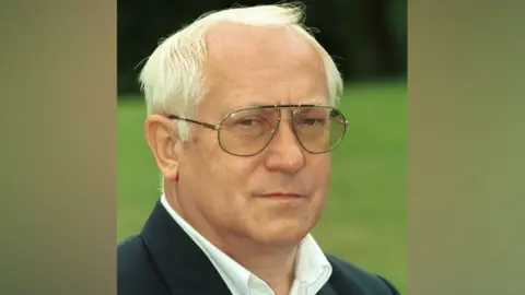 BBC Oleg Gordievsky looks at the camera, has short white hair and is wearing a white shirt under a dark suit jacket. There is a lawn in the background.