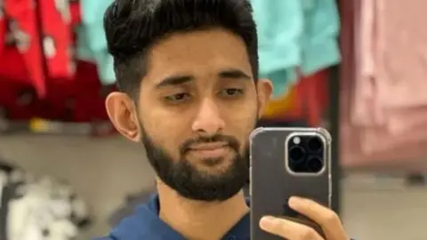 Man with dark hair and dark beard holds up a phone which he is looking into