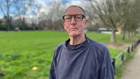 Martin Smart looking into camera next to a green park. He has short hair, dark rounded glasses and is wearing a navy jumper.