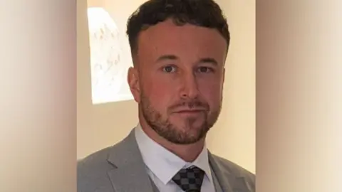 Man with a beard, wearing a white shirt and grey jacket looks into the camera