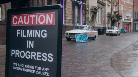 Northern Films Two Rover police cars parked up outside the former Bob Carver's chip shop in Trinity House Lane in Hull with a sign that reads "police slow" and a another sign that reads "caution, filming in progress, apologies for any inconvenience caused".
