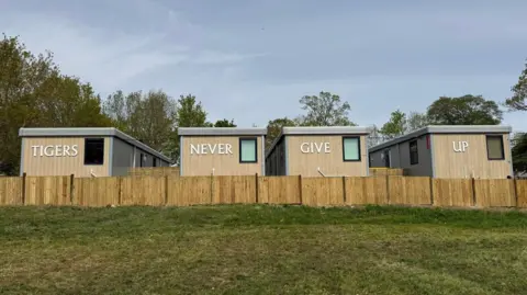 Four new prefabricated buildings in front of a field. Each one has writing on them which read: 'Tigers', 'Never', 'Give', and 'Up'.