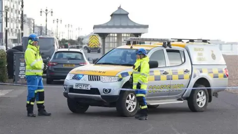 Eddie Mitchell Two members of the HM Coastguard stand behind a line of tape with a HM Coastguard vehicle on the beach