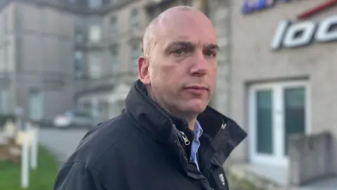 Louis Gardner looks into the camera wearing a black waterproof jacket and blue shirt 