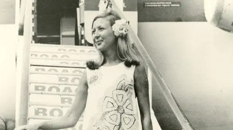 British Airways Heritage Centre A woman walks down the stairs off an aeroplane. She wears the paper dress design from 1967 and has a large flower in her hair, looking off to the left of the photo. 