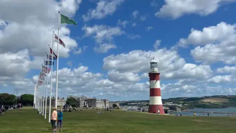 A row of flagpoles on Plymouth Hoe on the left with Smeaton's Tower, a red and white striped lighthouse on the right with the Hoe falling away to the sea.