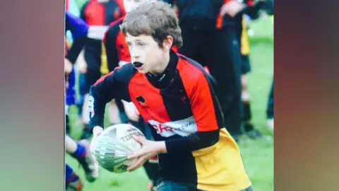 Old Coventrians/ Richard Allsopp Jake is running with ball-in-hand, with his gum shield showing in his mouth. He is wearing the Old Coventrians kit of black, red and yellow.