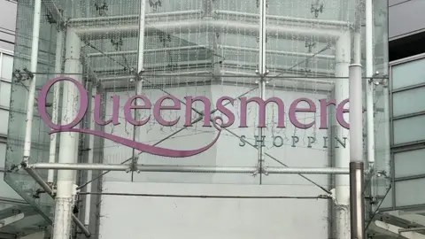 The sign of Queensmere shopping centre. 'Queensmere' is in a purple colour and 'shopping' is in a blue coral colour. 