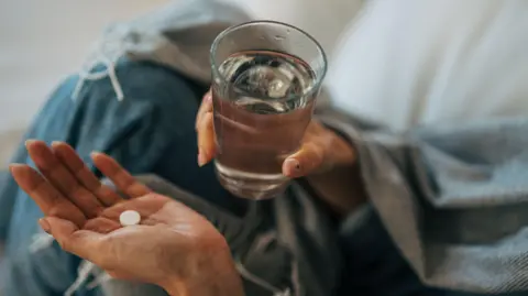 Getty Images A person holds a single white tablet in one palm and a glass of water in the other. They are wrapped in a grey blanket or shawl and wearing a denim jacket. The close-up shot focuses on the hands with the person's face out of frame.
