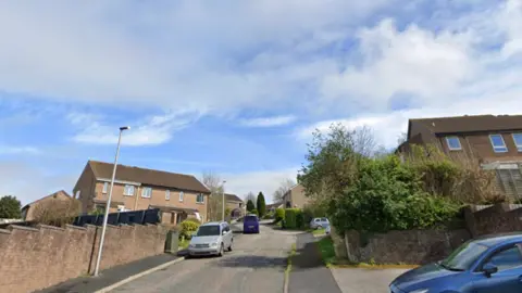 Image shows a residential street with semi detached houses and a road going up hill. Several cars are parked on the road and a blue car is parked on the right hand side of the frame in a parking space. The sky is blue with some white clouds. 
