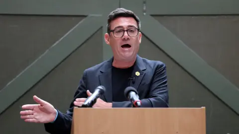 Reuters Andy Burnham, a man wearing a black t-shirt glasses and a black suit is standing at a lectern. There is a dark-coloured wall behind him. He is delivering a speech and there are two microphones visible pointing towards him.
