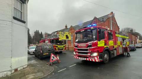 Kate Bradbrook/BBC Two fire engines are parked alongside and in front of a red brick building in the middle of the intersecting roads. The roads are wet and the sky grey.