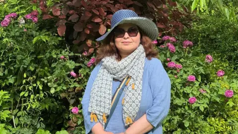 Saima Afzal Saima Afzal wears a floppy, blue sun hat, blue top, scarf and sunglasses as she stands in a garden in front of flowering shrubs.