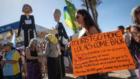 Getty Images A  woman holds a sign saying "tourists go home locals come back" during a protest in Tenerife, Canary Islands