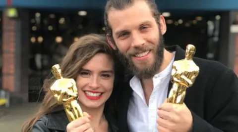 A woman and a man hold two gold Oscar statues in their hands.