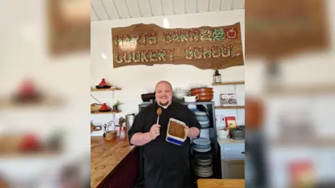 BBC Joe standing in his kitchen. He is wearing a black apron and is smiling to camera and holding a Tupperware box of the spread and a spoon with a mouthful on it. A large sign above his head says 'Harts Barn Cookery School'.