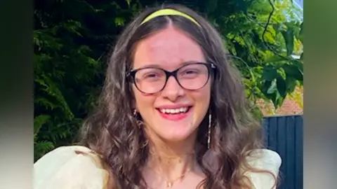 A headshot of Rosie, smiling. She has long brown, curly hair. She wears a yellow alice-band in her hair and black glasses. Her top is pale yellow and has puffy shoulders. 