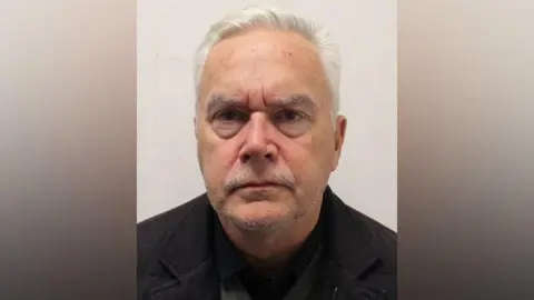 Metropolitan Police A custody image of a man with white hair and stubble on his face. He is wearing a black jacket and looking directly at the camera with a serious expression.