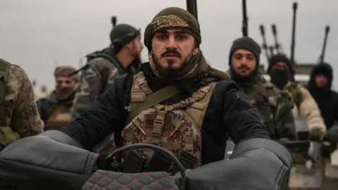 Syrian troops stand together as one soldier sits on a motorcycle, wearing tactical gear and surrounded by others carrying weapons.