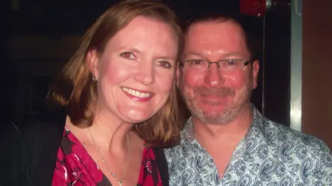 Handout A brown hair woman wearing a pink and black top. She is standing next to a man wearing a blue and white shirt. 