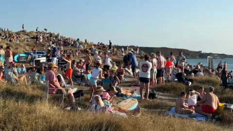 Lots of people of the sand and grassy coastline at Le Braye in Jersey. they are gathered on a hill to the left and down towards the sea on the right.