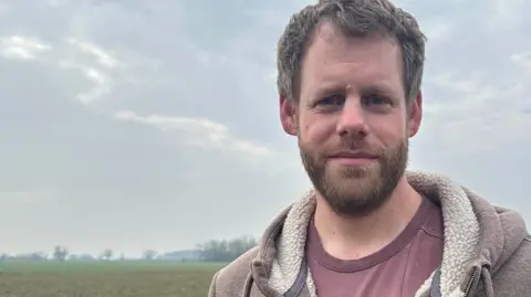 Jenny Kirk/BBC Chris Humphrey, with brown hair and a beard, wearing a dark pink tshirt and brown hoodie, standing in front of a field