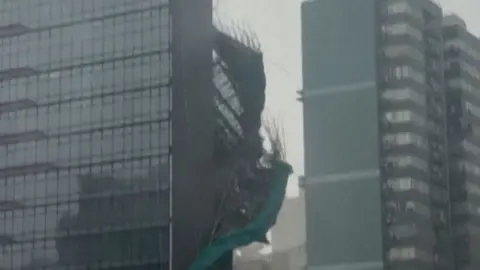 Bamboo and green plastic sheeting are seen falling down the side of a glass skyscraper.