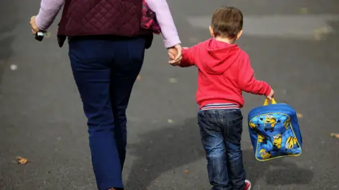 Brian Lawless/PA Wire A young boy wearing a red jumper and jeans is holding a woman's hand as they walk side by side. The small child is holding a blue Minions backpack in his other hand. The woman is wearing a purple jacket and jeans. She is holding keys in her other hand.