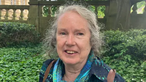 BBC/Grace Wood A woman with tied back grey hair wearing a colourful jacket and smiling at the camera