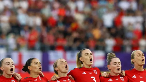 Reuters Six women in red Wales football shirts stood in a row with their arms round each other, heads raised and mouths open in song.