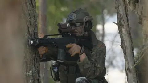 A soldier holds up a gun, while hiding amongst the trees, and aims somewhere out of frame