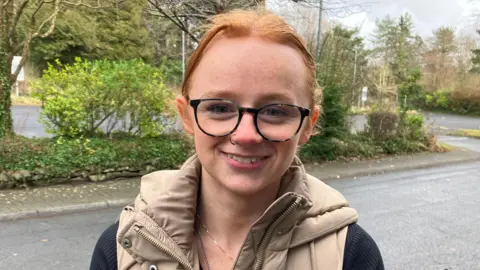 Erin, pictured on a pavement in front of a road with some bushes and trees. She has ginger hair tired back and wears tortoiseshell glasses. She smiles at the camera and is wearing a beige gilet and silver necklaces. 