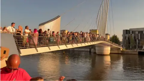 Image shows over a hundred people standing on a bridge at MediaCity, looking up into the sky
