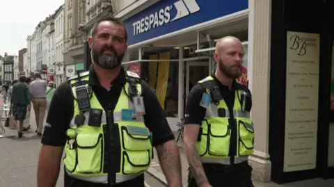 The image shows two men on patrol in Barnstaple's High Street, wearing hi-vis vests and walkie talkies.