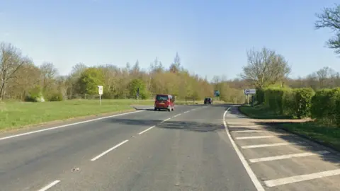 A road with a grassed area on the left and a low hedge on the right. Two cars in the left hand lane. A junction off to the left ahead with a sign on the opposite side of the road.
